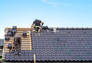 Two workers install black roofing tiles on a sloped house roof, with exposed wooden roof battens and stacks of tiles awaiting installation.