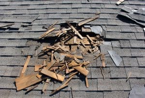 Damaged asphalt shingle roof with a large exposed section, broken wooden decking, and scattered roofing debris.
