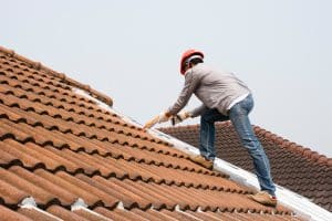 A roofer wearing a safety helmet works on a tiled roof, applying sealant along a roof joint to prevent water leakage.